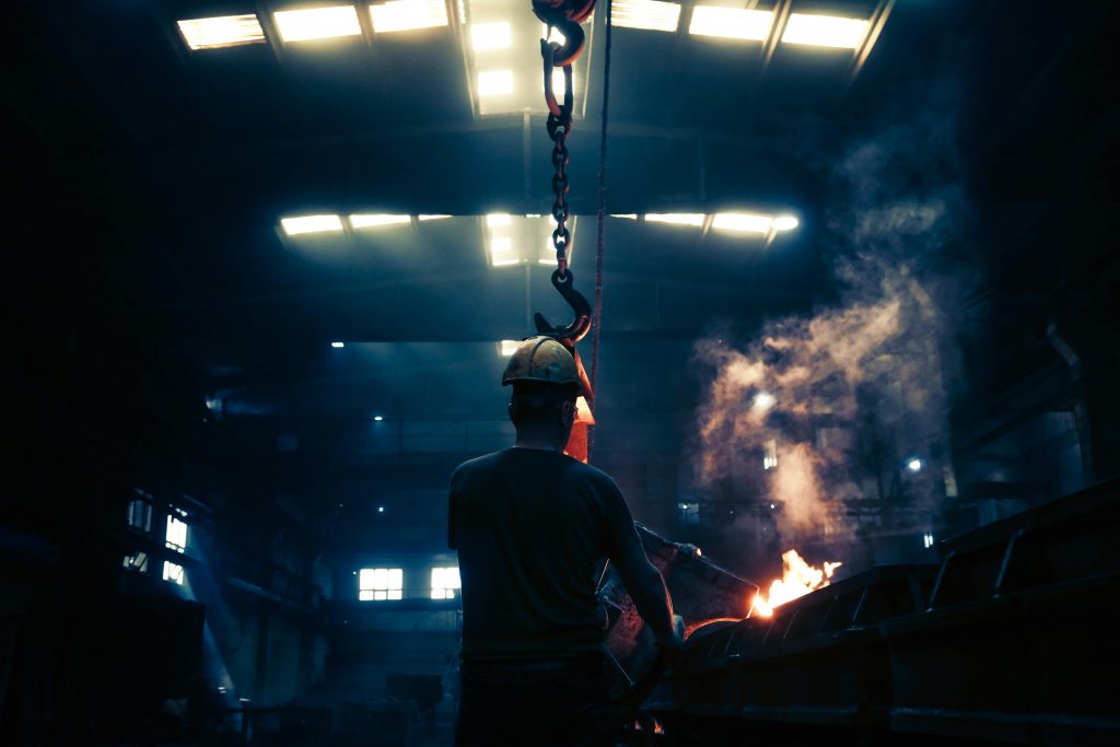 A worker in a hard hat operates machinery inside a large industrial factory with glowing flames.